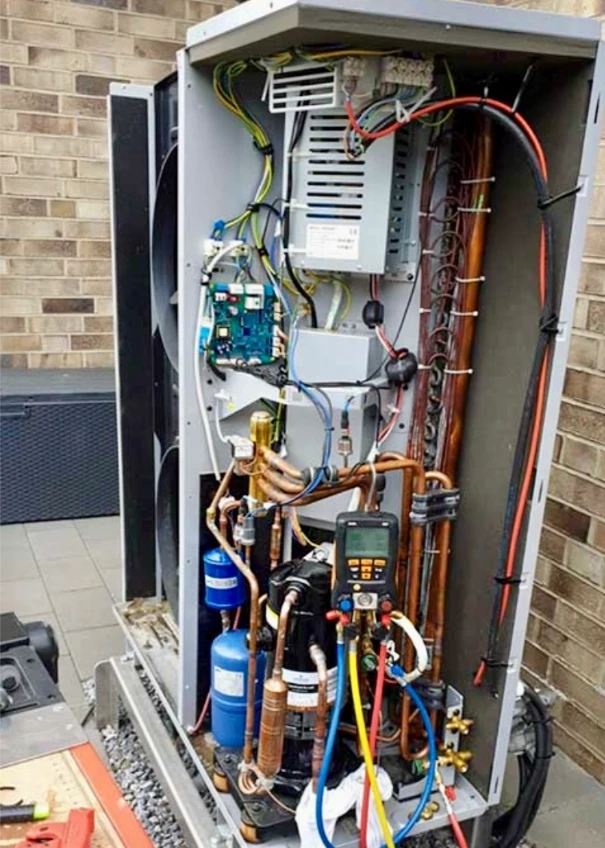 Widok wnętrza jednostki zewnętrznej pompy ciepła z rozłożonymi przewodami, modułem sterującym, rurami i manometrem